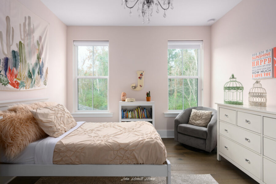 Bright modern bedroom with soft pink walls, large windows overlooking trees, and stylish decor including wall art, a cozy armchair, and elegant bedding, showcasing a comfortable home interior designed by John Merrill Homes.