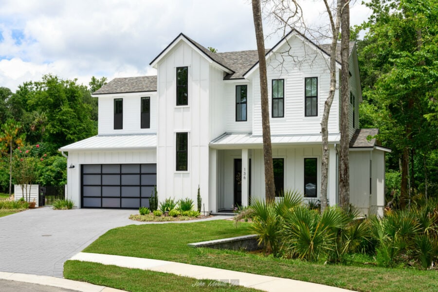 Modern white two-story home with a sleek black garage door, surrounded by lush greenery and landscaped garden, featuring large windows and contemporary architecture.