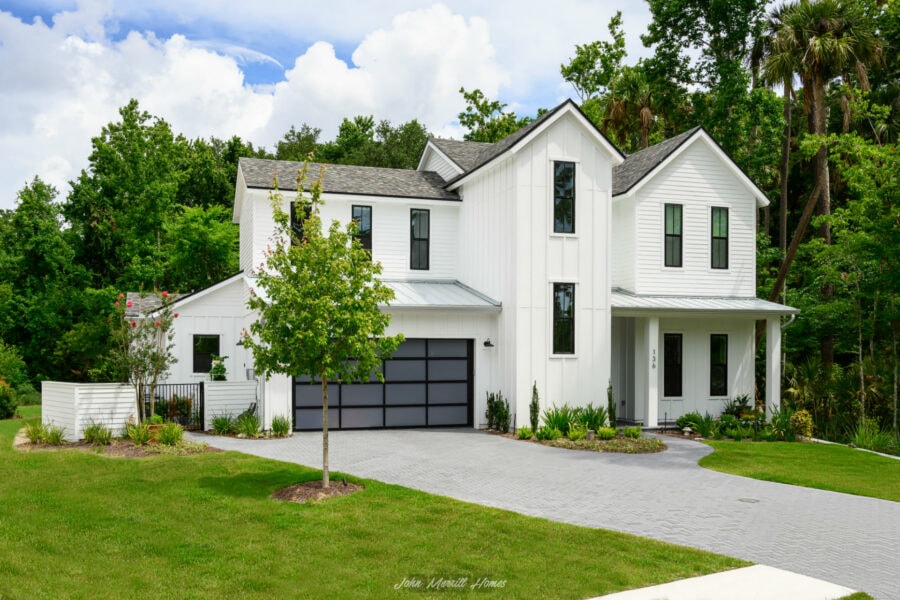 Modern white farmhouse-style home with black-framed windows, landscaped front yard, and paved driveway, surrounded by lush green trees and a bright blue sky.