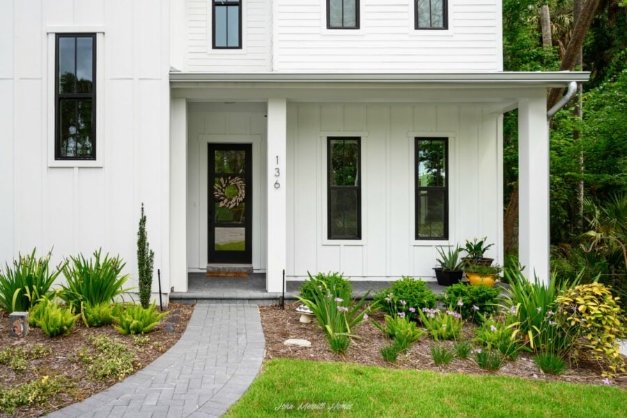 Front door of a modern white house with black trim, surrounded by lush green landscaping and a curved gray brick pathway, showcasing a welcoming and contemporary home exterior.