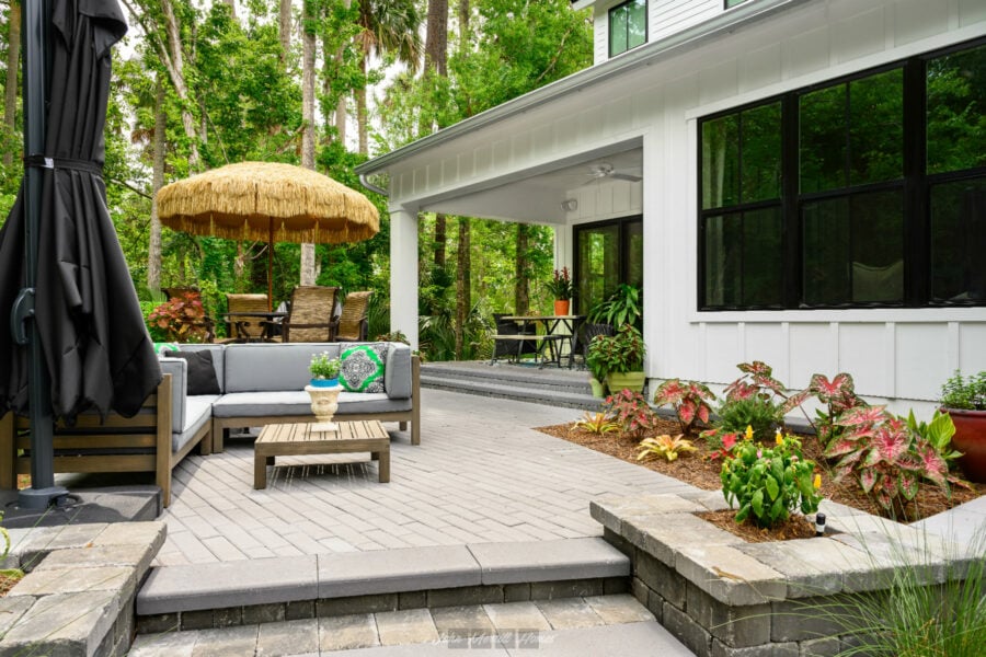 Bright outdoor patio with comfortable seating, potted plants, and a thatched umbrella, surrounded by lush green trees and landscaped garden, perfect for relaxing and entertaining.