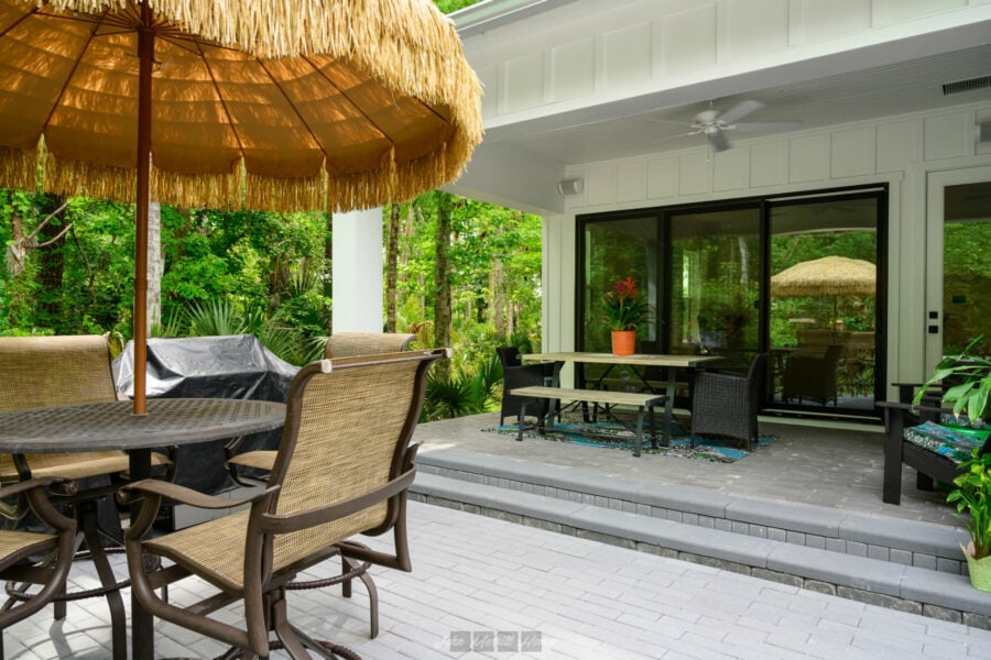 Covered outdoor patio with a round dining table, wicker chairs, a large straw umbrella, lush greenery in the background, and a sliding glass door leading into a modern house.