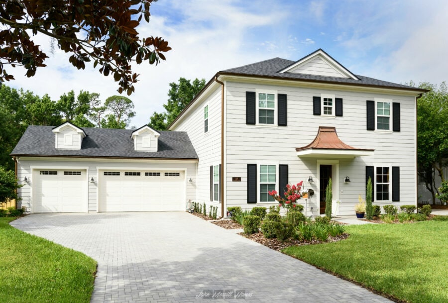 Modern, white two-story home with black shutters, a charming copper awning over the front door, and manicured landscaping, featuring a spacious paver driveway and lush green lawn.
