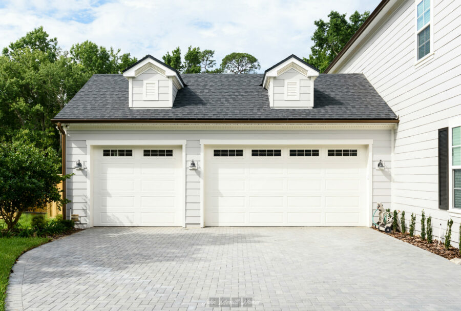 Two-car white garage doors with windows, part of a modern home's exterior with black shingle roof and white siding, surrounded by greenery and a paved driveway.