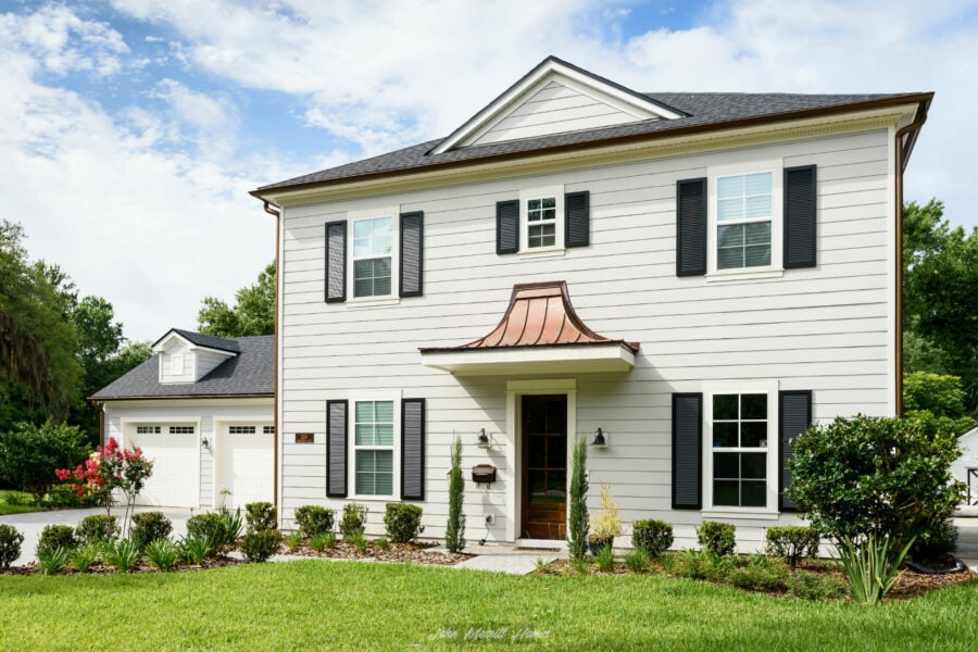 Modern two-story white house with black shutters, copper awning over front door, surrounded by lush green lawn and landscaped garden with bushes and flowering plants.