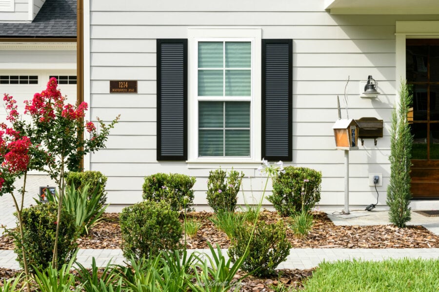 Freshly landscaped front yard with trimmed shrubs, blooming flowers, and a clean walkway leading to a modern home exterior with white siding and black shutters.