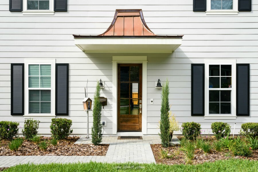Modern white house with black shutters, front door with glass panels, landscaped garden with small bushes and potted plants, cozy entrance, and elegant exterior design.