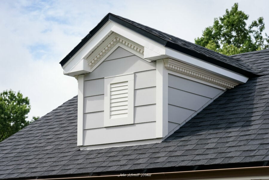Vent dormer with white siding, decorative trim, and a vent, set on a roof with dark gray shingles, surrounded by green trees and a partly cloudy sky.