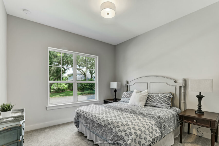 Bright bedroom with a large window, white walls, and a cozy bed with patterned bedding, complemented by black and white bedside lamps and a small plant in the corner, showcasing modern home interior design.