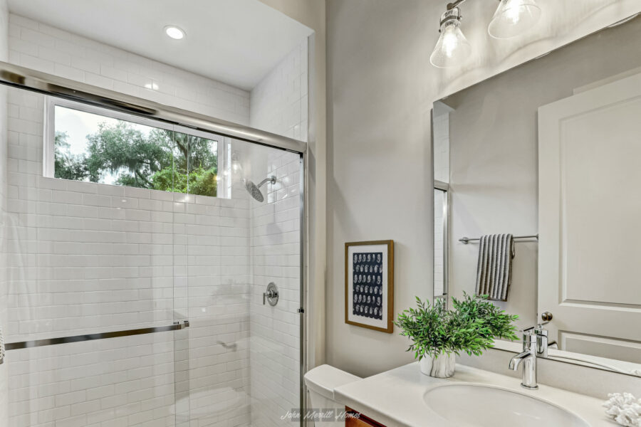 Bright modern bathroom with white tiled shower, large window, and stylish vanity with a mirror and decorative plant. Elegant fixtures and good lighting create a fresh, inviting space.