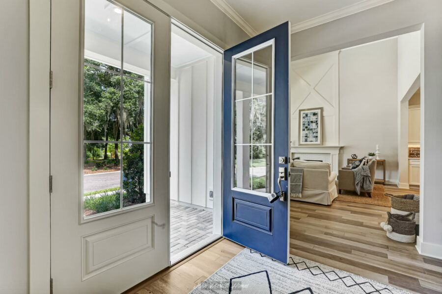 Bright blue front door opening into a welcoming, modern living room with large windows, white walls, and cozy furnishings, overlooking a green outdoor space, creating an inviting home entrance.