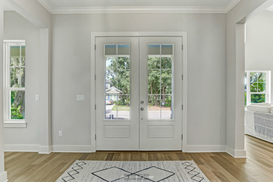 Bright white double glass doors leading outside, surrounded by natural light, with a view of a lush green residential neighborhood.
