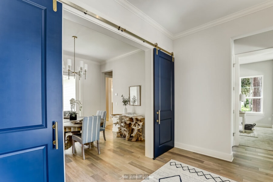 Vibrant blue sliding barn doors opening into a modern, bright dining area with a wooden table, striped upholstered chairs, and elegant chandelier. Soft natural light illuminates the tidy, stylish space.