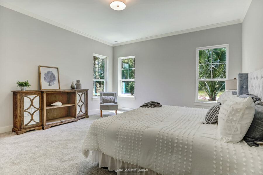 Spacious bedroom featuring a large bed with textured white bedding, a wooden sideboard with decorative accents, and three windows providing natural light and greenery view.
