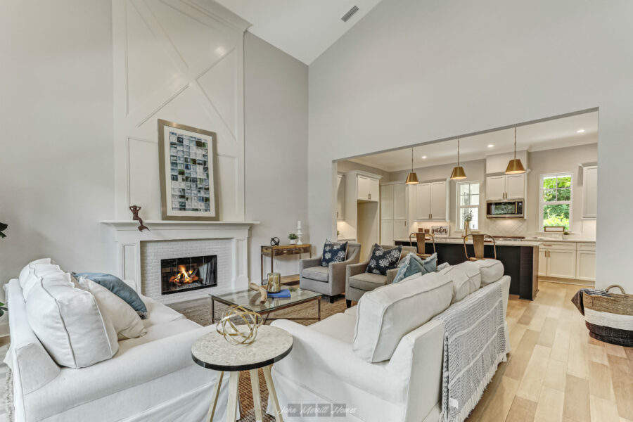 Modern living room with a cozy fireplace, light-colored sofas, and contemporary decor, open to the kitchen with white cabinetry, pendant lighting, and large windows for natural light.