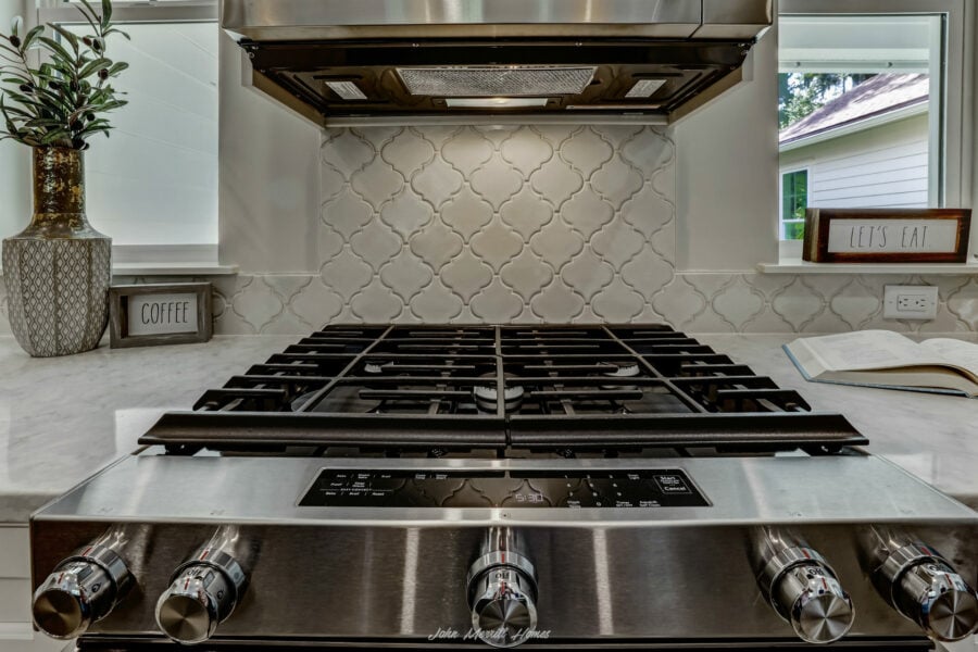 Stainless steel gas stove with four burners on a white marble countertop, decorated with potted plants and signs reading "COFFEE" and "LET'S EAT" in a modern kitchen.