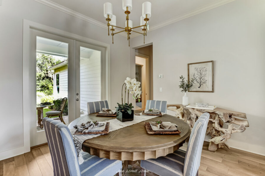 Elegant dining room with a rustic wood table, stylish upholstered chairs, and a modern chandelier, opening to an outdoor patio with lush greenery, showcasing contemporary home design.