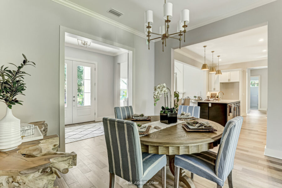 Bright dining area with a round wooden table, upholstered chairs, and elegant decor, opening to a sunny entryway and modern kitchen in a spacious, freshly designed home.