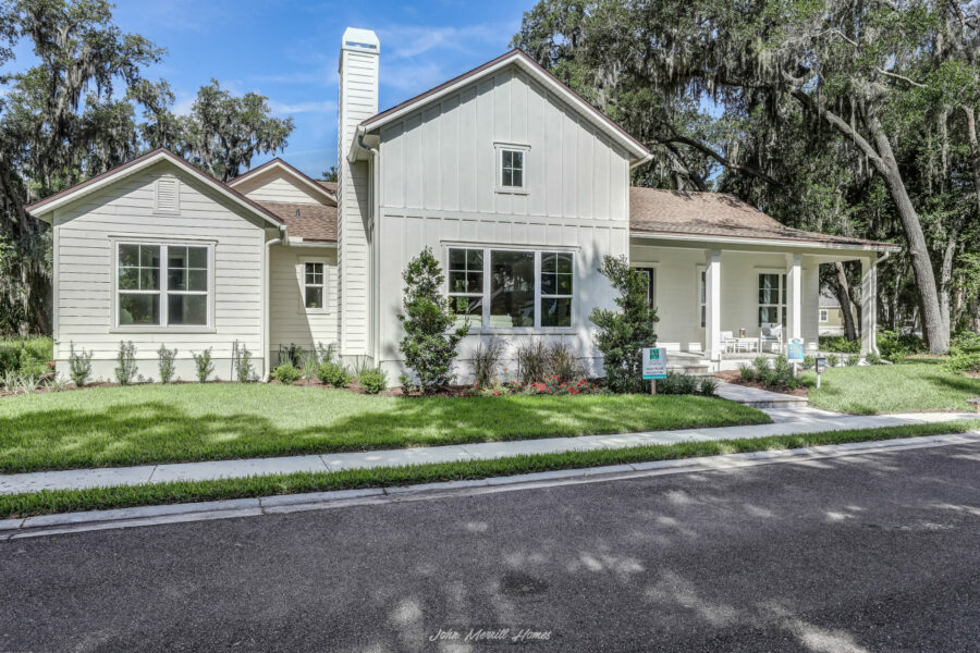 Spacious white modern farmhouse-style home with a large front porch, surrounded by well-manicured lawn and landscaped garden, set against a backdrop of mature trees.
