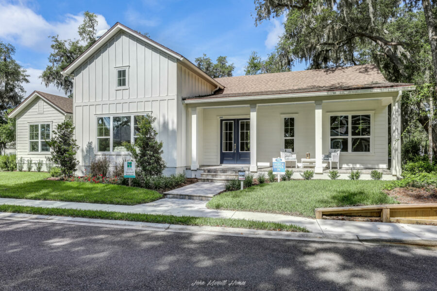 Modern white farmhouse-style home with a welcoming front porch, black double doors, landscaped yard, and mature trees in the background, highlighting new construction by John Merrill Homes.