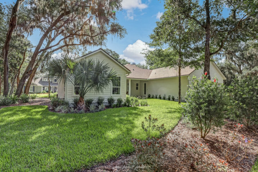 Lush green backyard with mature trees, trimmed grass, and landscaped plants surrounding a white house with siding and large windows, creating a peaceful suburban setting.