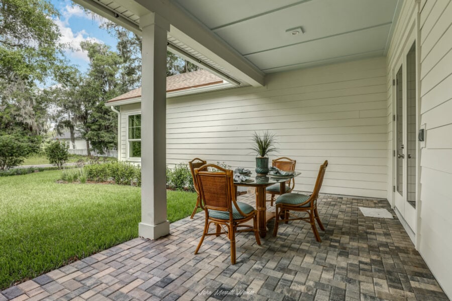 A covered outdoor patio with a glass-top dining table, four wicker chairs with cushions, lush green lawn, and white siding house exterior, ideal for relaxing and outdoor entertaining.