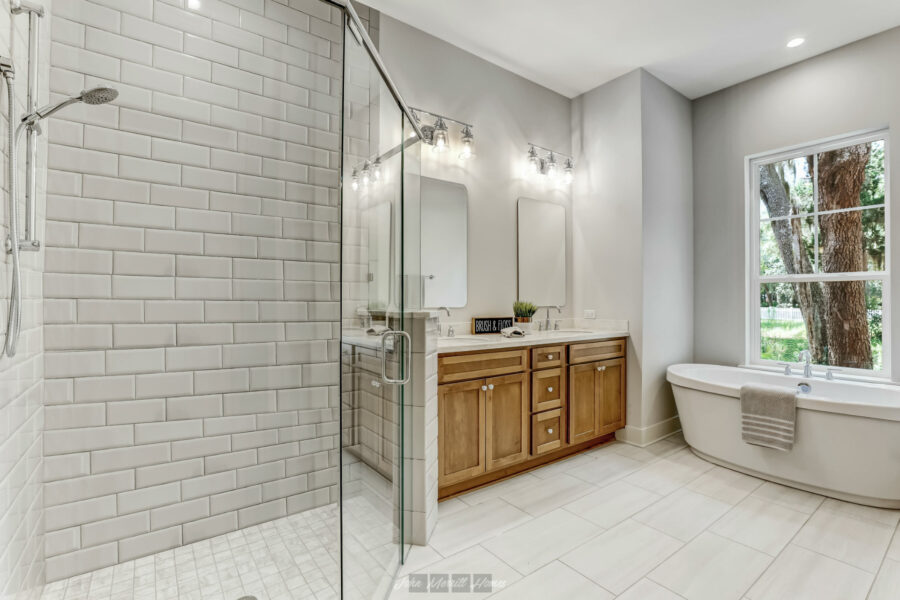 Modern bathroom with a white tiled walk-in shower, a wooden vanity with dual sinks and mirrors, a large window overlooking nature, and a freestanding bathtub for relaxation.