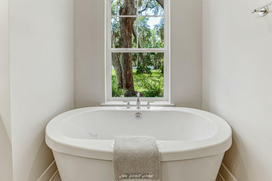 Spacious white bathtub in a bright bathroom with a large window showing lush green trees outside.