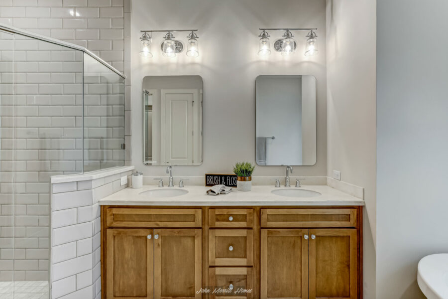 Bright bathroom vanity area with dual sinks, wooden cabinetry, large mirrors, modern lighting, and a walk-in shower with white subway tiles.