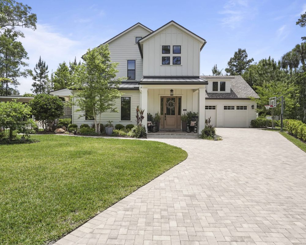 Modern white farmhouse-style home with a landscaped front yard, paved driveway, and a basketball hoop, surrounded by lush trees and greenery under a clear blue sky.