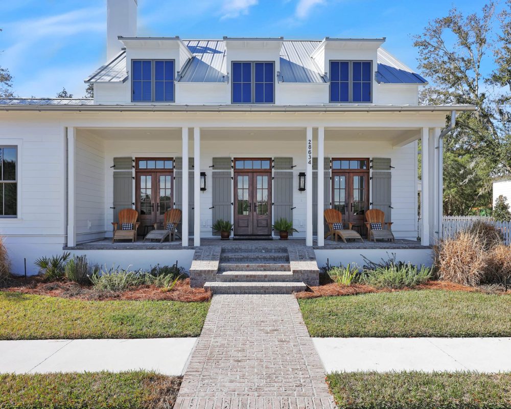 Modern white farmhouse with a spacious front porch featuring wooden chairs, planters, and brick steps, set against a bright blue sky with well-maintained landscaping and mature trees.