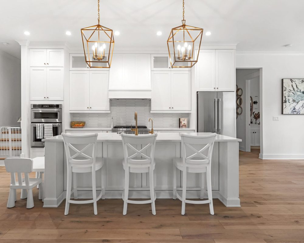 Modern white kitchen with an island, gold pendant lighting, and seating; spacious and bright interior with minimalistic decor and wooden flooring.
