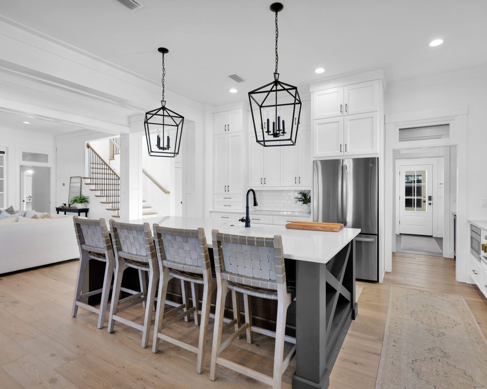 Modern white kitchen with a large island, black pendant lighting, stainless steel appliances, and hardwood flooring, part of a spacious open-concept home by John Merrill Homes.