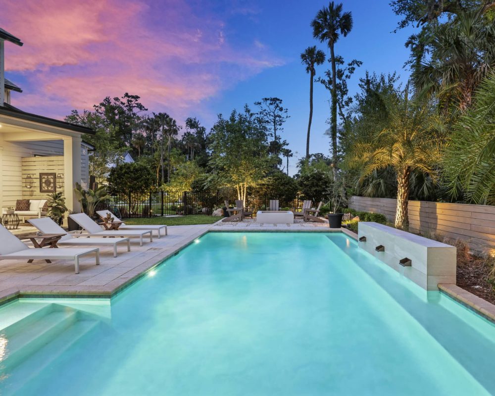 Luxurious outdoor swimming pool at twilight with lounge chairs, lush tropical landscaping, cozy seating area, and vibrant sunset sky, showcasing elegant backyard design by John Merrill Homes.