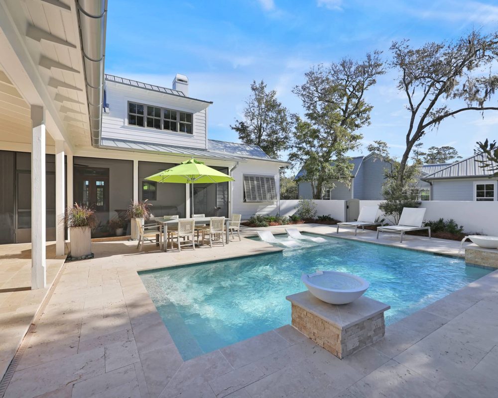 Luxurious outdoor swimming pool with lounge chairs, a dining area with a table and umbrella, surrounded by a modern patio, trees, and a stylish house with large windows and white siding.