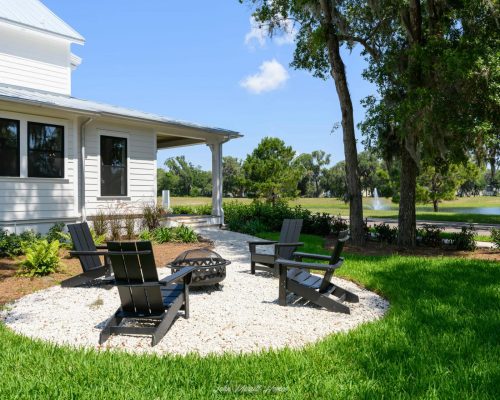 Fire pit area with black Adirondack chairs on white gravel, surrounded by lush green lawn and trees, overlooking a pond with a fountain at John Merrill Homes.