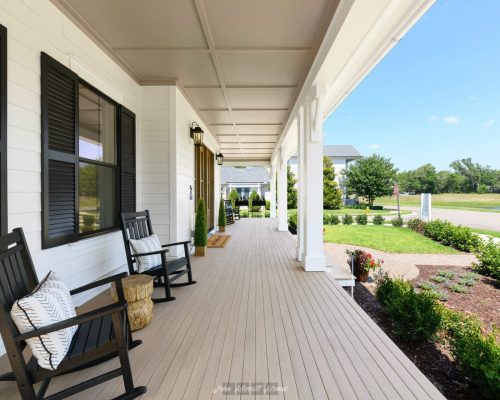 Porch with black rocking chairs, decorative pillows, and potted plants outside a white house with black shutters, overlooking a lush green lawn and street on a sunny day.