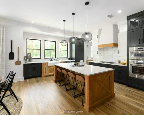Spacious modern kitchen with black and white cabinetry, a large central island with wooden accents, pendant lighting, and large windows providing natural light.