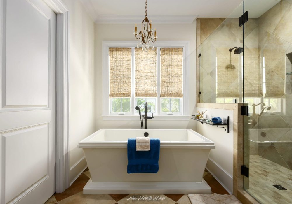 Luxurious bathroom featuring a white bathtub, large window with woven shades, elegant chandelier, and a glass-enclosed shower with beige tiles, creating a spa-like retreat.