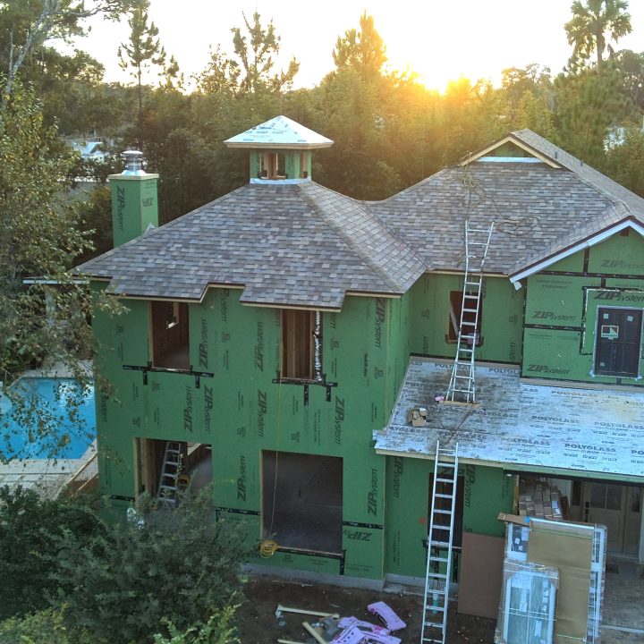 New two-story house under construction with green sheathing, surrounded by trees, during sunset, with a worker on the roof and construction materials nearby.