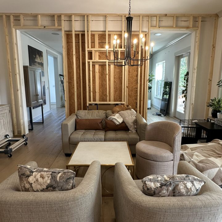 Modern living room with unfinished wooden wall framing, beige sofa, armchairs, and a black chandelier, showcasing a contemporary interior design under construction.