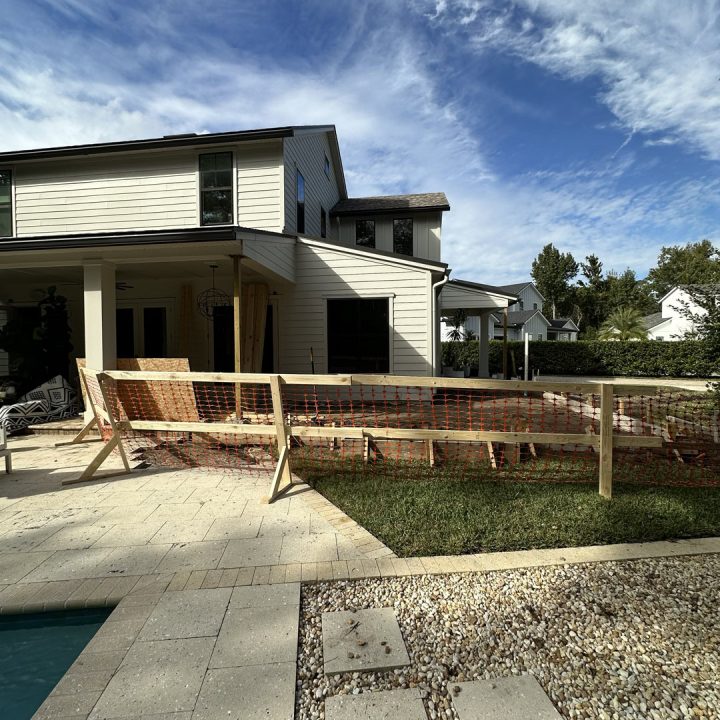 Modern two-story house under construction with a spacious backyard, clear blue sky, and construction fencing in the foreground, showcasing contemporary architecture and ongoing building work.