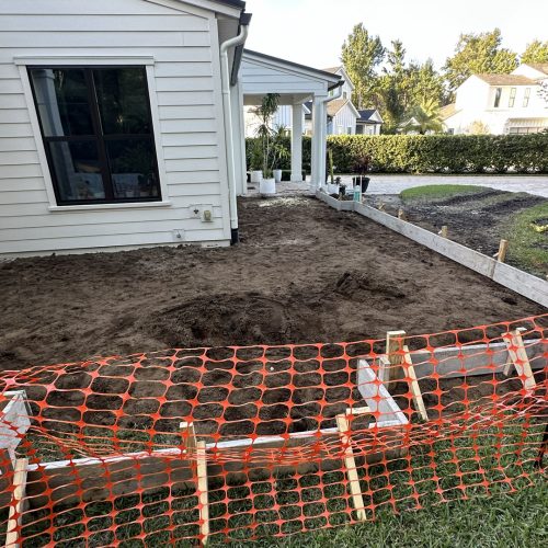 New construction home foundation preparation with dirt work in progress, white house siding, and landscaping underway at a residential property.