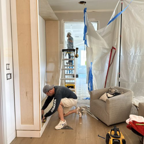Newly constructed home interior under renovation with workers installing built-in shelves, showcasing modern living space design and construction progress.