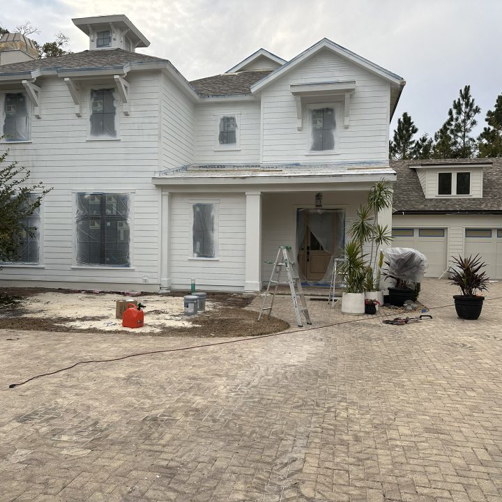Newly constructed two-story white house with a front porch under construction, surrounded by scaffolding and tools, featuring multiple windows, a side garage, and a paved driveway, with trees in the background.