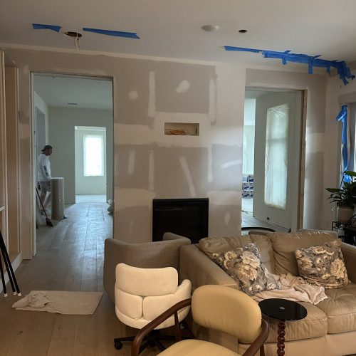 Bright living room under renovation with drywall, construction materials, and a worker visible in the background. Blue painter's tape and protective coverings on windows indicate ongoing interior updates.