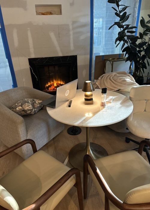 Cozy living room with a modern white fireplace, a round marble table, comfortable beige armchair, and stylish black and gold accents, showcasing a warm, inviting interior of a new home under construction.