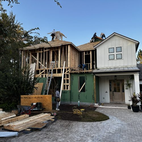 New home construction in progress with workers actively building the roof and frame structure of a modern house, featuring a front porch with potted plants and a paved driveway.