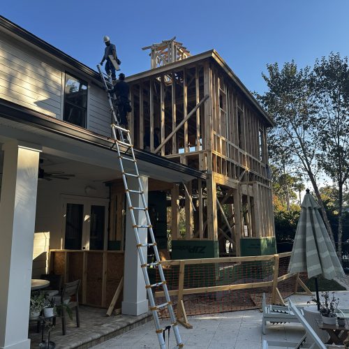 Newly constructed multi-story residential building in progress with workers installing framing; the backyard features outdoor furniture, a pool, and lush trees under a clear blue sky.