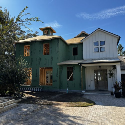 New home under construction with white exterior siding, multiple windows, and a paved driveway, featuring modern farmhouse design, surrounded by trees and a clear blue sky.